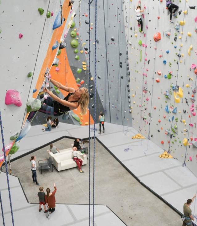 A photo looking down on the indoor climbing gym of CLIMB Bentonville. Lead ropes dangle in the foreground, with a climber on a rope contemplating their next move up the wall.