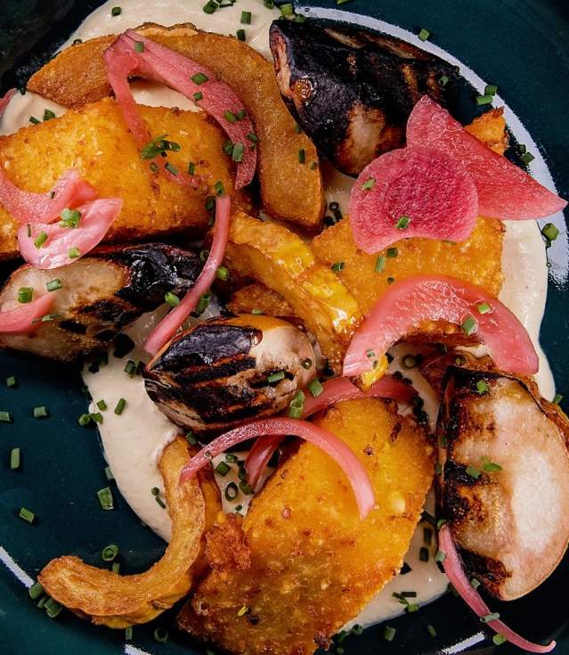 A vibrant citrus salad with pink radish slices, roasted delicata squash, and grilled onions served on a dark green bowl, embodying the fresh, seasonal fare of Conifer restaurant.