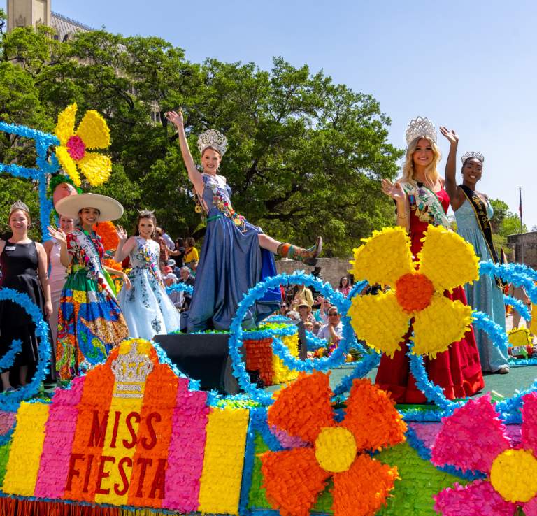 Women dressed in gowns and crowns on a colorful Fiesta float in front of the Alamo.