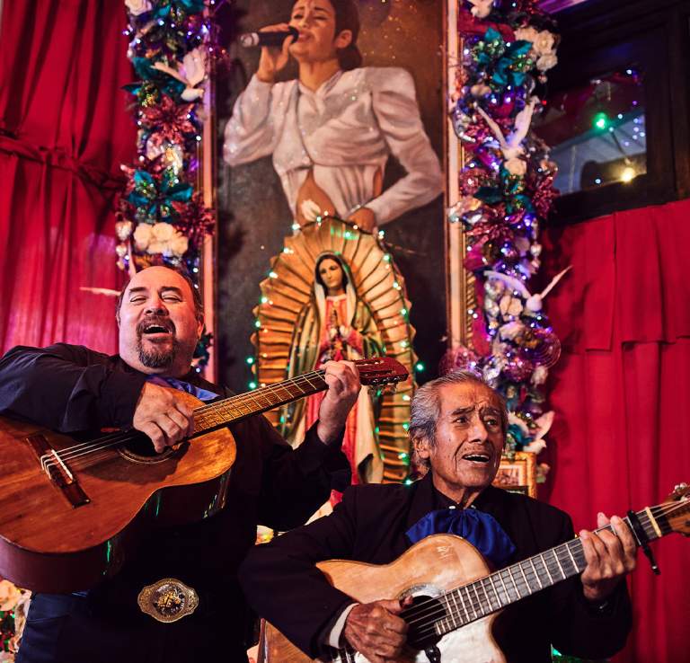 Two mariachi players in front of a painting of the Tejano artist Selena