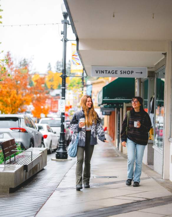 Two people stroll past shops on a rainy autumn day in downtown Nanaimo, holding coffee as colourful fall trees line the street.