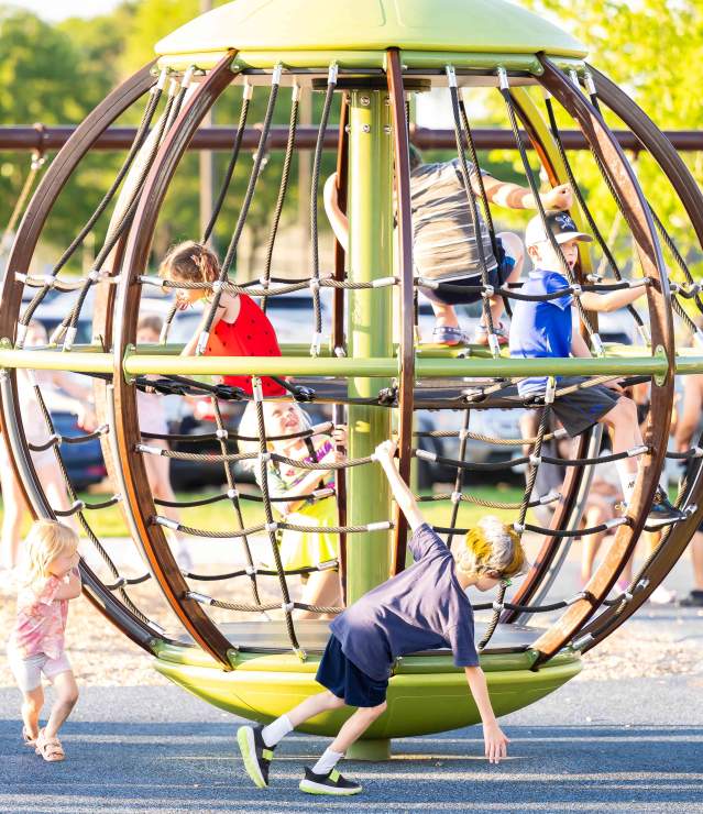 Kids playing at park on a spinning net carousel / rope sphere with teens swinging in background.