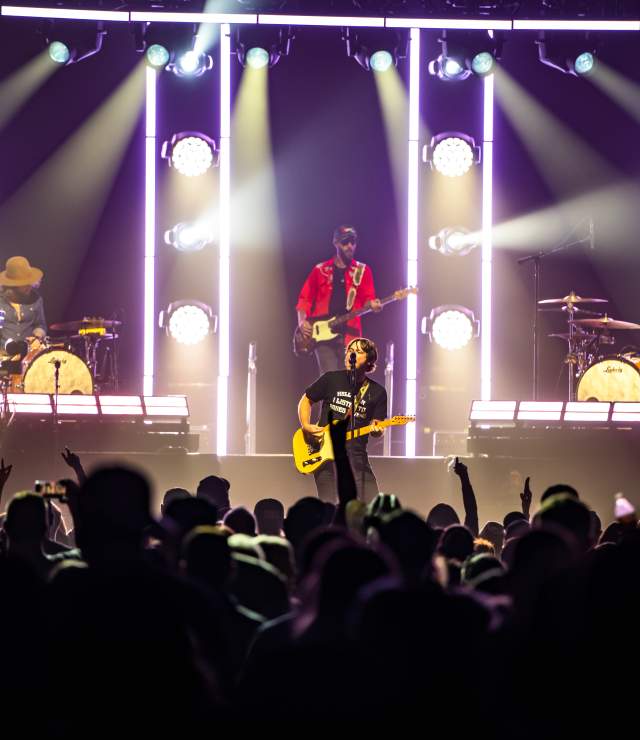 A live music concert featuring a full band performing on stage, with dramatic vertical light bars and colorful spotlights illuminating the scene. The lead singer stands center stage with a yellow electric guitar, flanked by guitarists, a bassist in a red shirt, and two drummers. The crowd in the foreground is silhouetted, many raising hands and taking photos, contributing to the vibrant, high-energy atmosphere of the show.