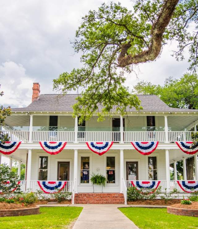 two story white historic home and museum with red, white, and, blue flags draped on the balcony, and trees