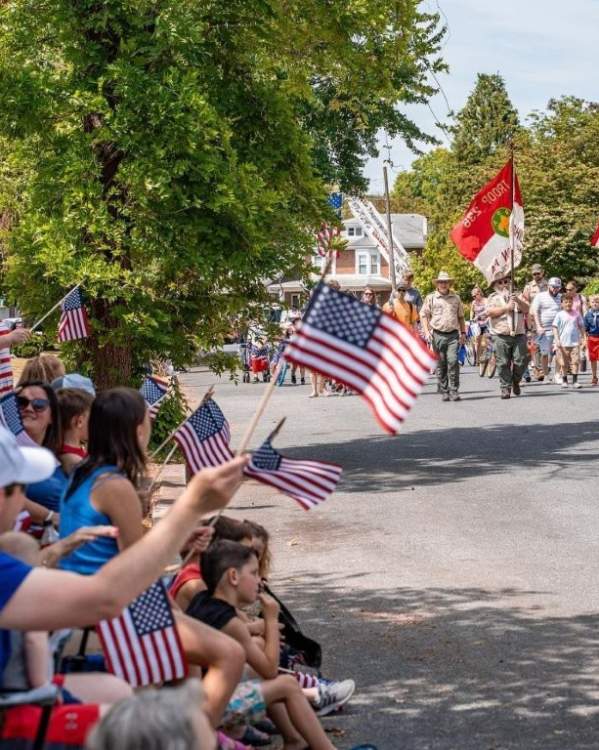 Spectators line a tree-lined neighborhood street waving American flags as a patriotic parade passes by. Scouts carrying flags lead the procession while families watch from lawn chairs and the curb under a sunny summer sky.