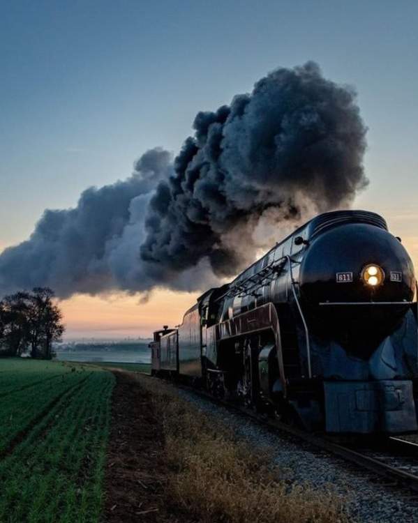A black steam locomotive travels through open farmland at sunset, billowing a thick plume of dark smoke into the sky. The train's bright headlight illuminates the tracks ahead as it passes fields and a small cluster of trees, with the warm colors of the setting sun glowing along the horizon.