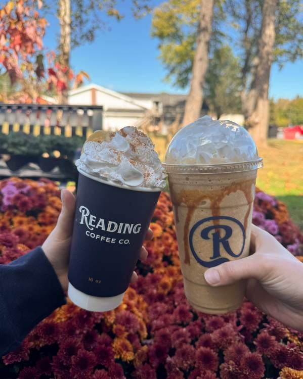 Two hands hold a hot latte topped with whipped cream and cinnamon and a caramel blended coffee topped with whipped cream from Reading Coffee Co. in front of colorful fall mums on a sunny autumn day.