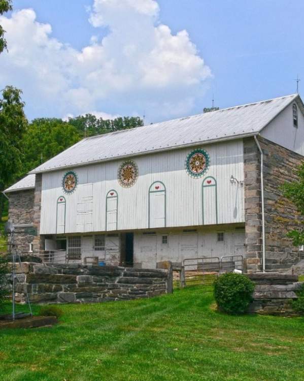 A large traditional Pennsylvania bank barn with white wooden siding, stone end walls, and a silver metal roof sits on a gently sloping grassy property. Decorative painted hex signs adorn the front of the barn, while stone retaining walls, fencing, and mature trees surround the historic rural scene beneath a blue sky with large white clouds.