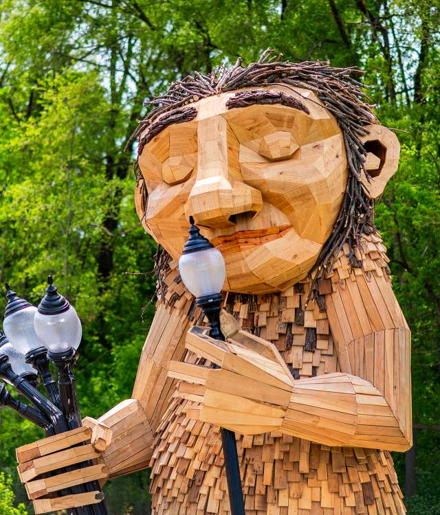 A large wooden sculpture of 'Mama Rosa,' a Thomas Dambo troll installation in Wauwatosa. The friendly giant figure is constructed from reclaimed wood pieces in natural tones, featuring a carved face with a gentle smile and twig-like hair made from bundled branches. The troll holds a bouquet of old-fashioned street lamps in one hand while lifting one lamp to her nose. The sculpture is set against a lush green forest backdrop with dense tree canopy, creating a whimsical scene that blends art with nature.