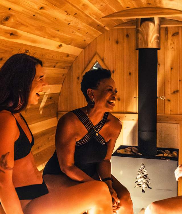 Two women in swimwear sit inside a warm, wood-paneled sauna at Heat Haven in Wauwatosa, smiling and talking near a small heater that glows softly in the center of the room.