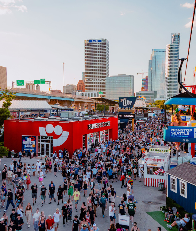 A large crowd moves through the bustling Summerfest grounds in Milwaukee during the day, with the downtown skyline visible in the background. A red building with the Summerfest smiley-face logo serves as the festival store. Above, colorful gondolas on the Skyglider transport people across the venue. The scene is filled with booths, food stands, and signage under a clear sky.