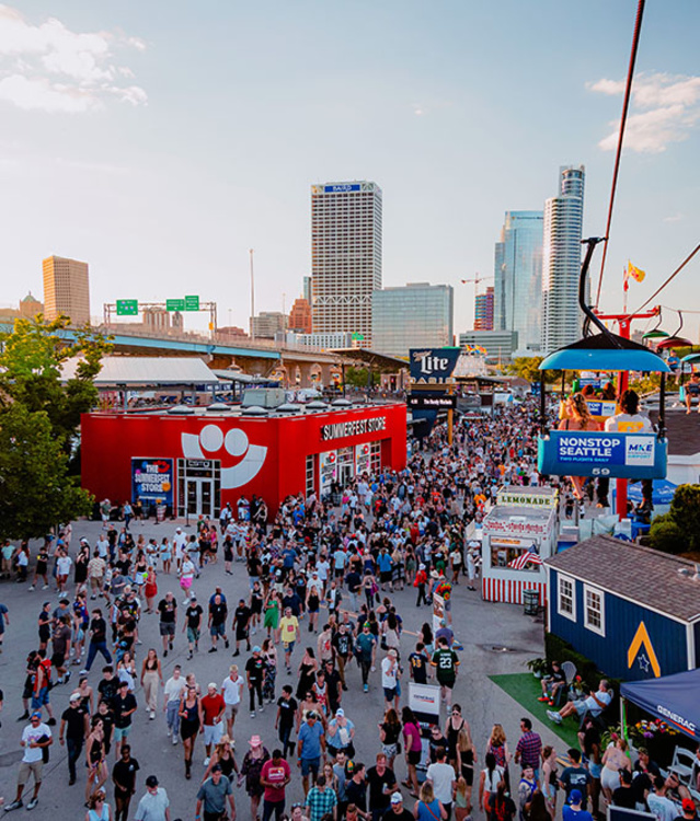 A large crowd moves through the bustling Summerfest grounds in Milwaukee during the day, with the downtown skyline visible in the background. A red building with the Summerfest smiley-face logo serves as the festival store. Above, colorful gondolas on the Skyglider transport people across the venue. The scene is filled with booths, food stands, and signage under a clear sky.
