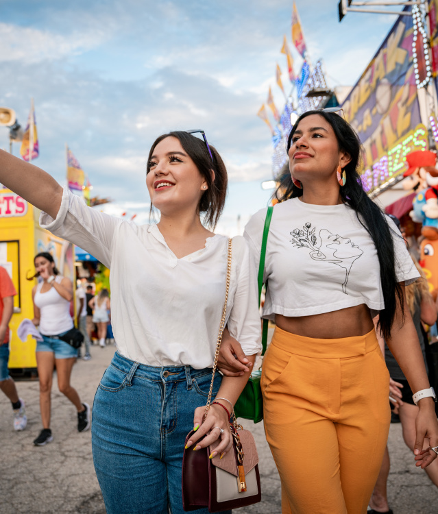 Two young women walk arm-in-arm through the Wisconsin State Fair, with one pointing excitedly ahead. Carnival rides, game booths, and crowds create a festive atmosphere in the background.