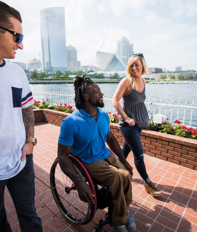 Three friends, including one using a wheelchair, wander along Milwaukee’s lakefront with the skyline in view.