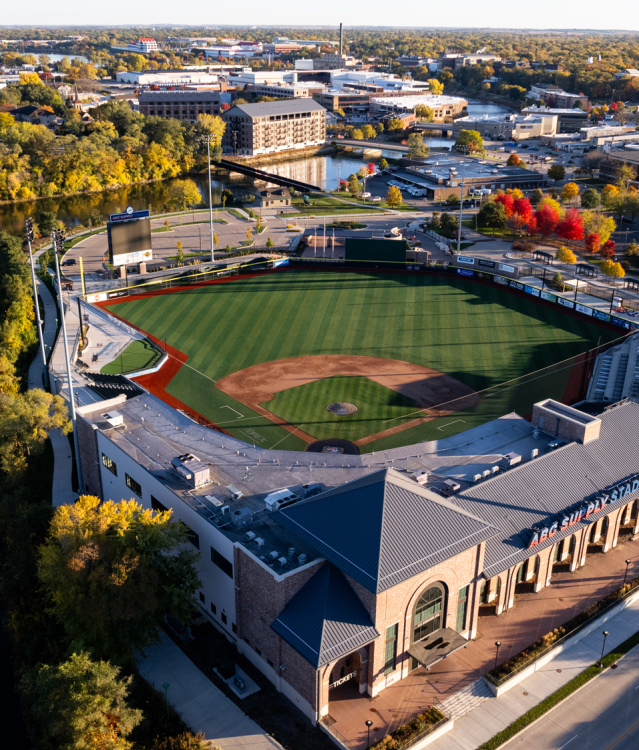 An aerial view of ABC Supply Stadium beside a winding river, surrounded by colorful autumn foliage and the city skyline. The baseball field sits at the center of the scene, with nearby streets, buildings, and a water tower visible under clear skies.