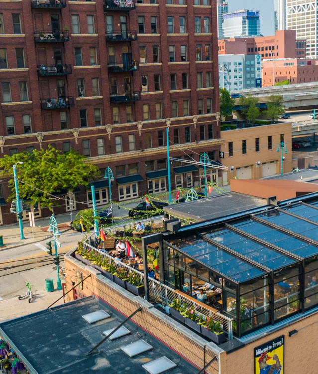 The Lux Skyhaus at Cafe Benelux, a glass greenhouse rooftop dining space in Milwaukee, overlooks a pedestrian plaza with teal lamp posts. The transparent structure reveals lush greenery and diners inside, surrounded by historic red brick buildings and downtown Milwaukee's skyline.