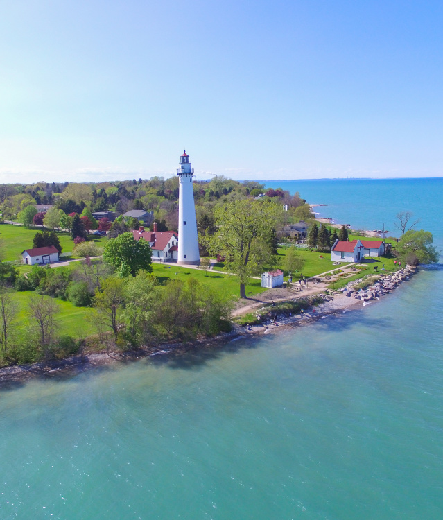Aerial view of a white lighthouse and keeper’s buildings on a green lakeside point, surrounded by trees and lawns along the shoreline of Lake Michigan. Clear blue water stretches into the distance beneath a bright sky, with the lighthouse standing prominently above the landscape.
