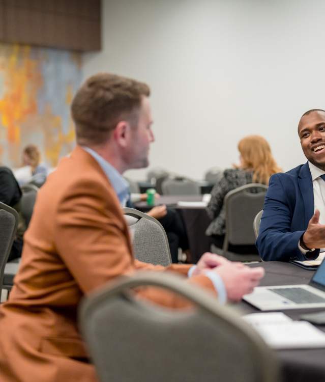 Two business professionals conversing at a conference table - one in an orange blazer with back to camera, the other in a navy suit smiling and gesturing while speaking. Multiple attendees visible in background conference room setting.