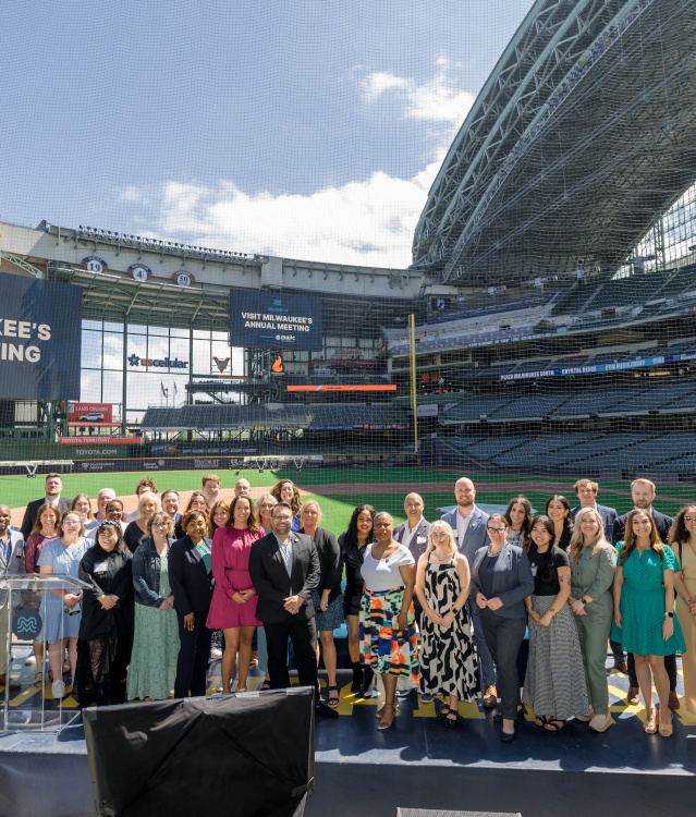 Group photo of attendees at Visit Milwaukee's Annual Meeting, posed on a stage at American Family Field (Milwaukee Brewers stadium) with stadium seating and banners visible in the background.