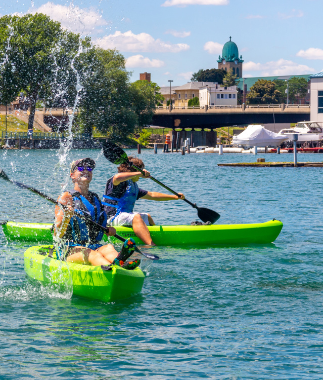 Two people paddle bright green kayaks on a sunny harbor, splashing water as they kayak across blue water. Trees, a bridge, boats, and waterfront buildings line the shore in the background beneath a clear sky.