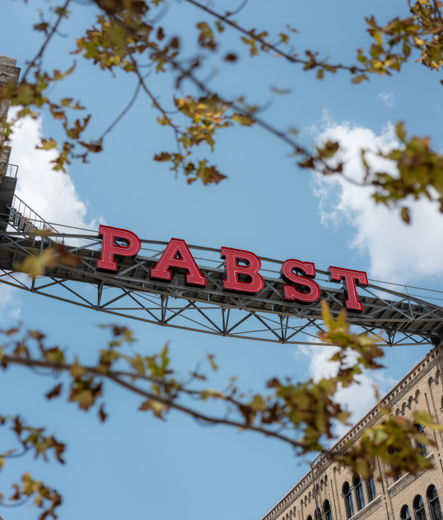 The iconic red Pabst sign stretches between historic brick buildings in Milwaukee, framed by fall leaves against a blue sky.