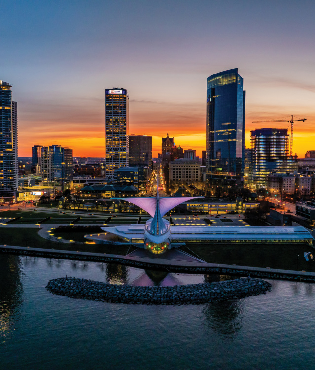 An aerial view of Milwaukee's lakefront at sunset features the Milwaukee Art Museum and the downtown skyline glowing against a colorful orange and blue sky. The calm waters of Lake Michigan reflect the evening light, highlighting the city's iconic waterfront.