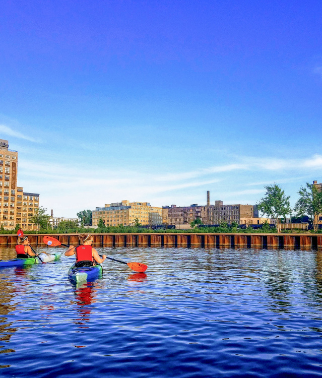Two kayakers on the Menomonee River with calm waters and and urban backdrop