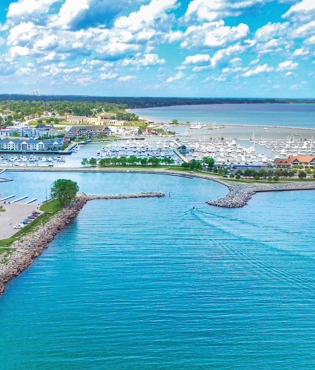 An aerial view of Reefpoint Marina along the Lake Michigan shoreline, featuring rows of docked boats, a protected harbor, and vibrant blue water. Waterfront buildings, green spaces, and a lakeside cityscape stretch beneath a sky filled with scattered clouds.