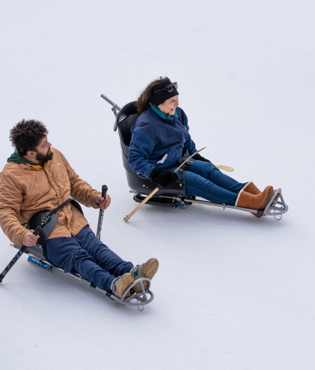 wo people ride seated ice sleds, using short poles to push themselves across an ice rink while others skate nearby.