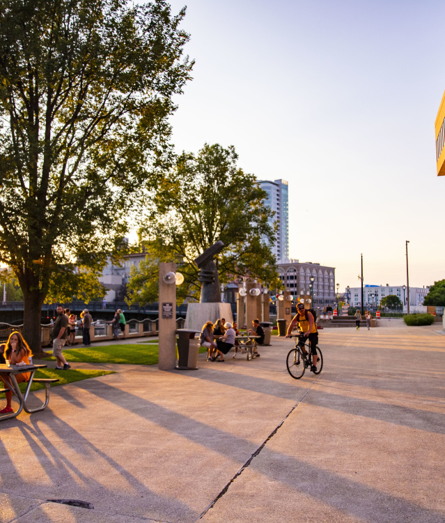 A vibrant urban plaza scene at the Marcus Performing Arts Center RiverWalk during golden hour, featuring people relaxing at picnic tables on green grass while others cycle and gather on the wide concrete plaza. The warm sunlight filters through mature trees, casting long shadows across the space.