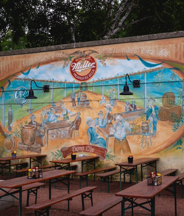Wooden picnic tables line the South Shore Beer Garden in Milwaukee, with a colorful Miller beer mural depicting historic brewery scenes on the wall behind them.