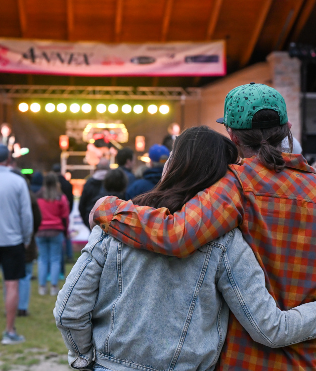 A couple stands arm in arm watching a live outdoor concert from the audience. Stage lights glow beneath a covered pavilion as community members gather on the lawn to enjoy the performance.