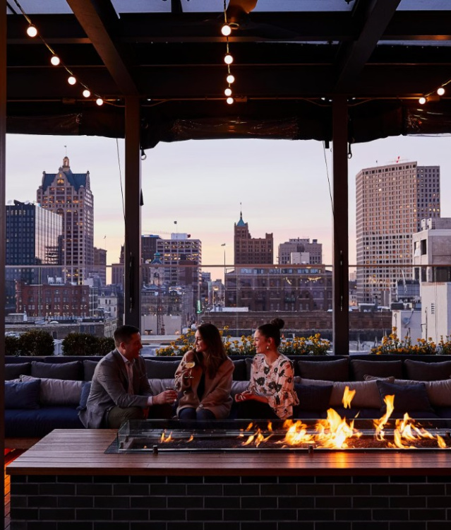 Three people sitting around a rooftop fire pit at sunset, enjoying drinks with a view of downtown Milwaukee’s skyline at The Outsider rooftop bar.