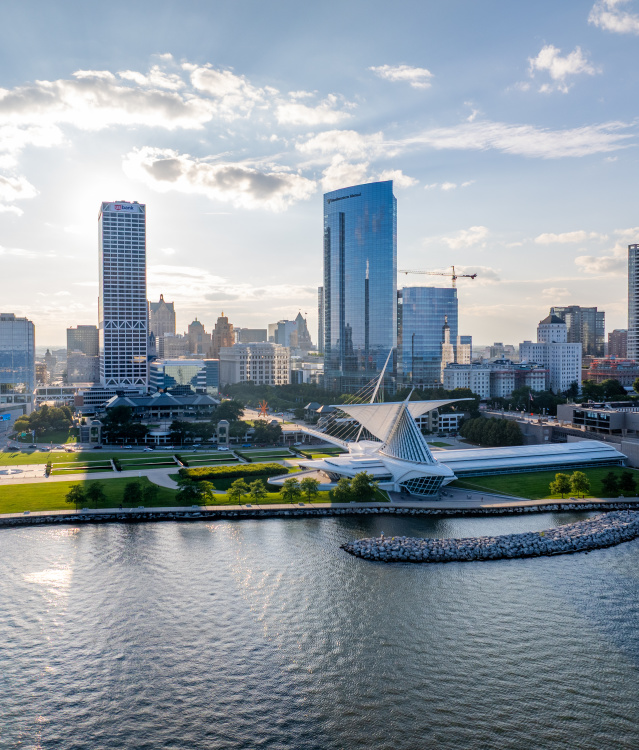An aerial view of Milwaukee's lakefront showcases the Milwaukee Art Museum, Discovery World, and downtown skyline along the shore of Lake Michigan. Sunlight reflects off the water as the city's modern architecture and waterfront parks stretch into the distance.
