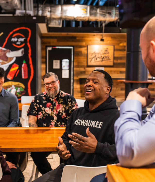 LeRoy Butler is wearing a black Milwaukee hoodie speaks animatedly while seated at a table inside 3rd Street Market Hall, with several people listening and smiling around him