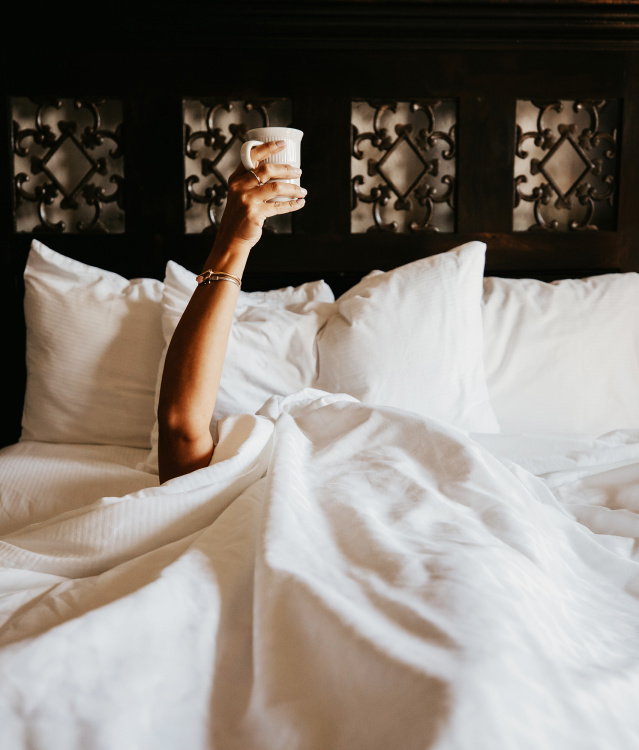 person in bed under covers with only the arm and coffee mug showing