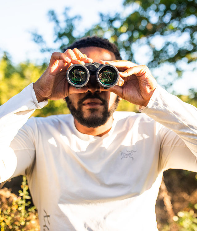 A man in a white long-sleeved shirt holds binoculars up to his eyes while standing outdoors in a sunny, tree-filled setting with green foliage and dappled sunlight in the background.