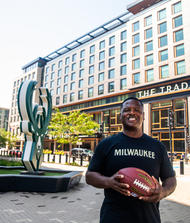 LeRoy Butler smiles while wearing a black ‘Milwaukee’ T-shirt holds a football in an outdoor plaza, with The Trade Hotel and a large deer sculpture visible behind him in Deer District.