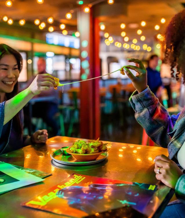 two women sitting at a table in a dim, string-light-lit bar pulling apart a cheese curd to stretch the cheese inside