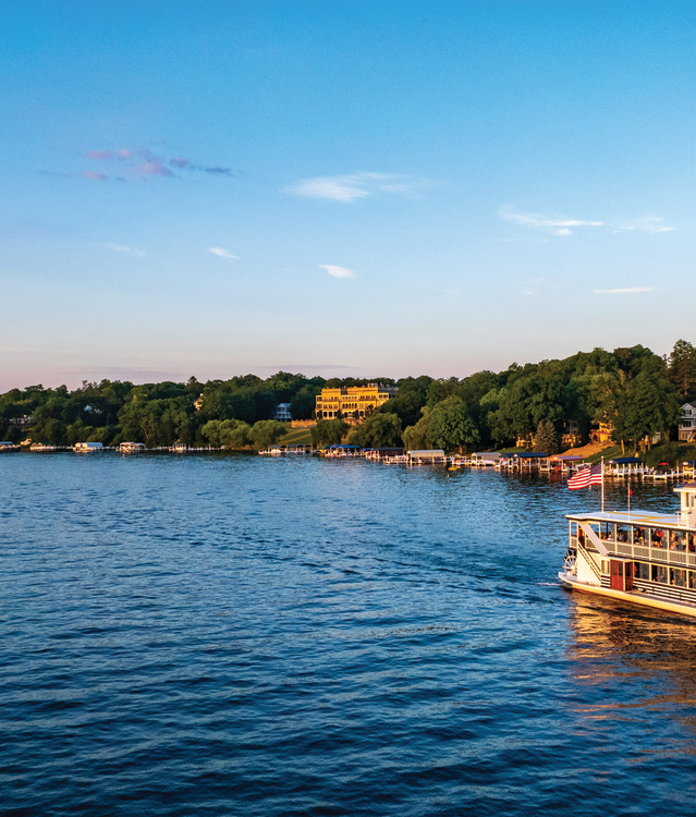 A tour boat cruises across a calm lake at sunset, with passengers aboard and shoreline homes nestled among trees in the background. Warm golden light reflects off the water, creating a scenic and relaxing lakeside view.