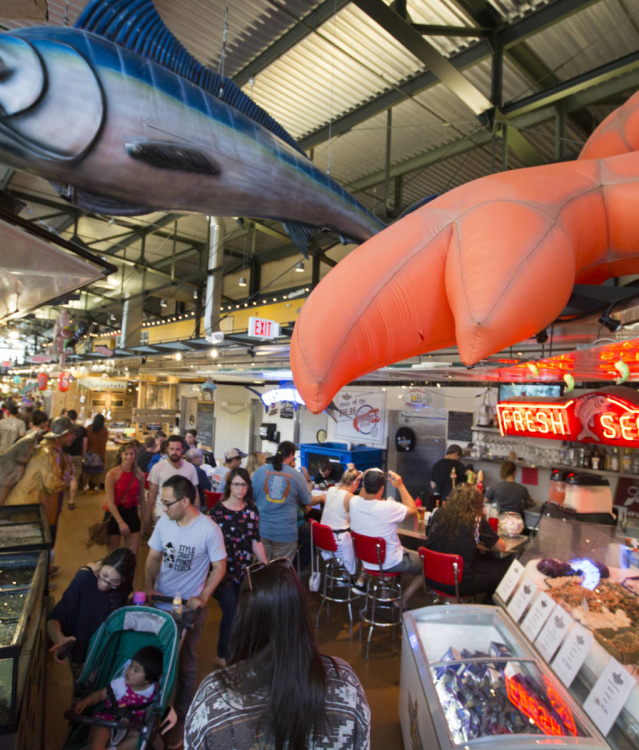 A bustling indoor seafood market filled with people browsing and dining, featuring large hanging fish decorations, bright neon “Fresh Seafood” signs, and displays of fresh catches on ice.