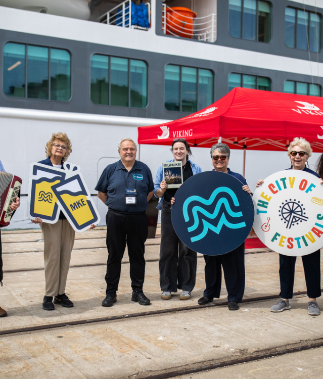 A welcoming group stands dockside in front of a cruise ship and red canopy, holding Milwaukee-themed signs including “The City of Festivals” and a stylized “MKE” logo, while an accordion player adds a festive touch.