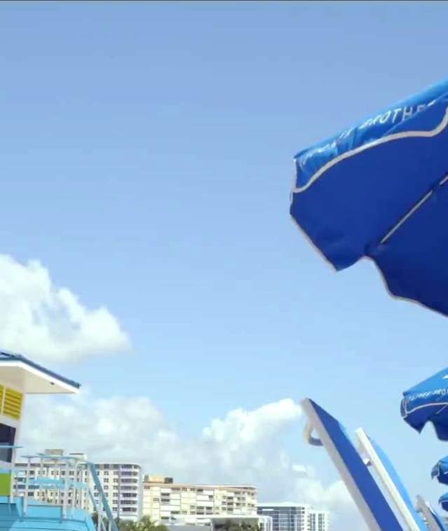 The tops of a line of open blue beach umbrellas with the Pompano Beach logo on them. A colorful lifeguard stand is visible in the background.