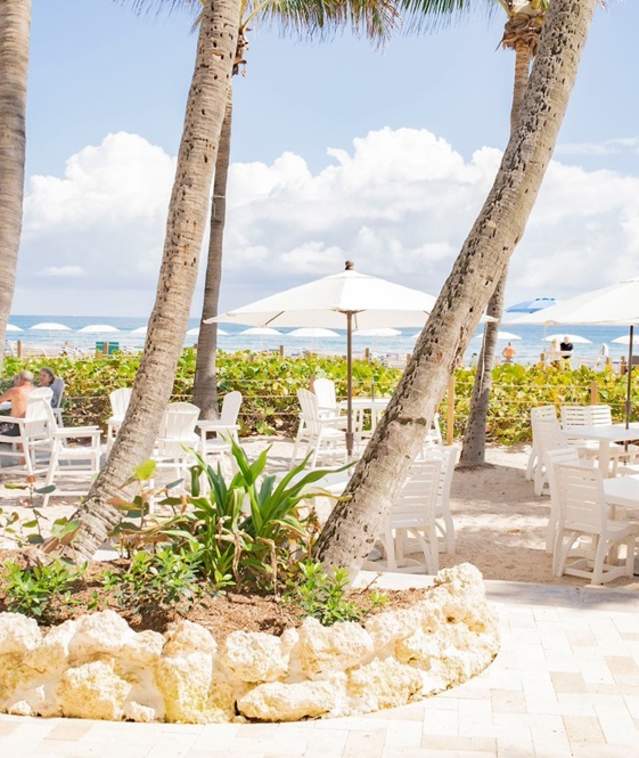 Several tables and chairs with umbrella set in the sand with an ocean view, for table service.