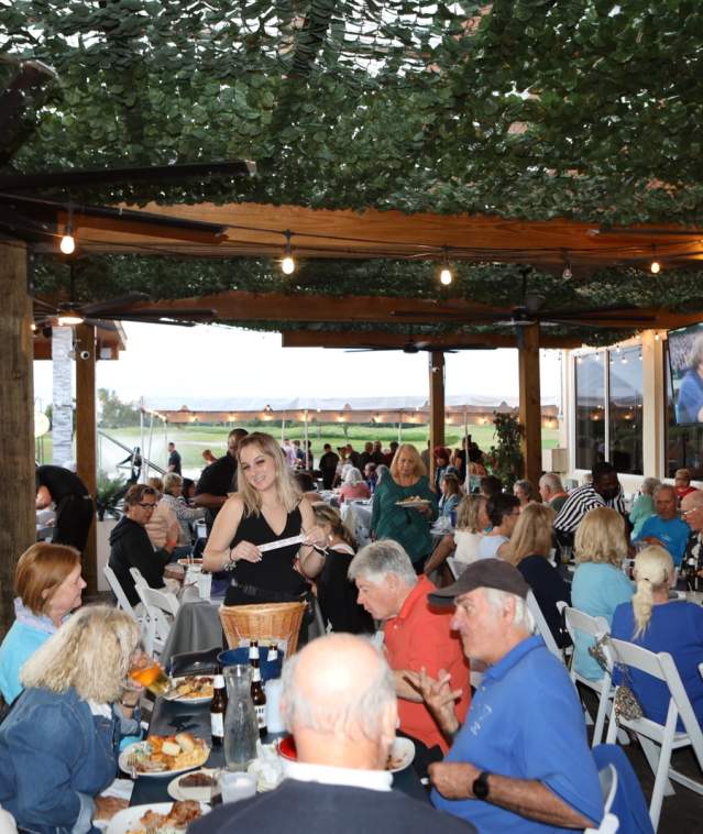 a covered patio with many tables filled with diners watching a large outdoor tv screen