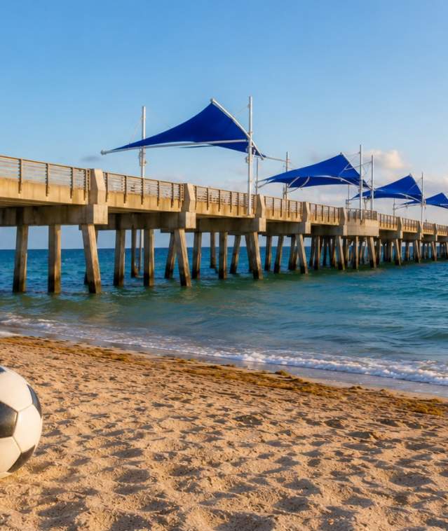 A soccer ball on the beach with the Pompano Beach Pier in the background.