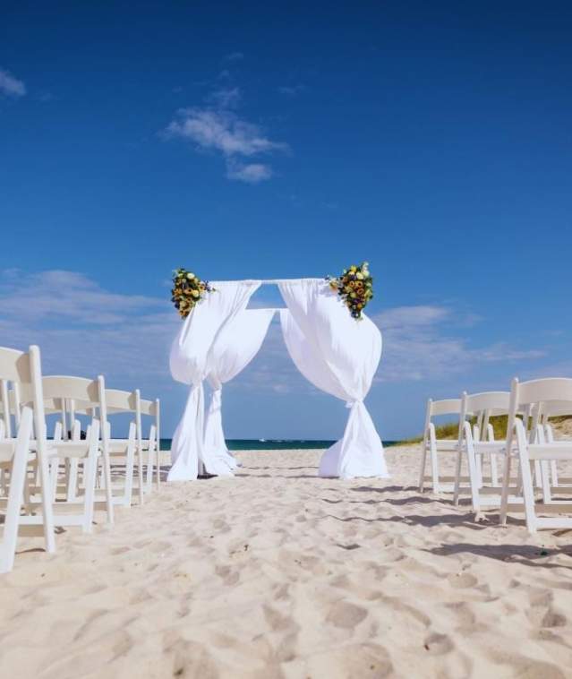 A wedding arch set up on the beach with white folding chairs lined up on either side of an aisle leading tot he arch