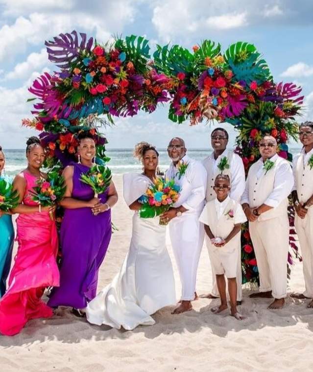 A bridal party posed in front of a very colorful arch on a beach with the ocean in the background. The bridesmaids are dressed in bright colors of the rainbow and the groomsmen, bride and groom are all dressed in white in Pompano Beach.