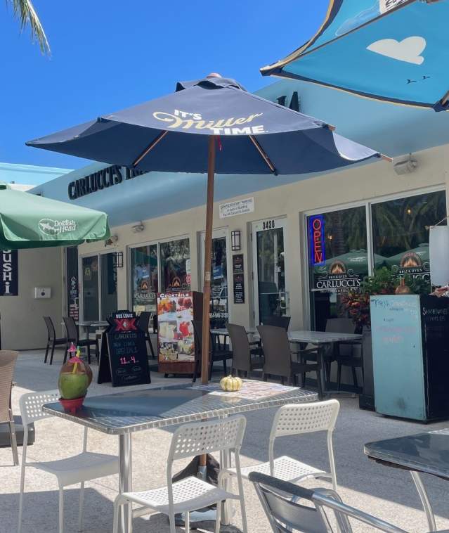 A strip of restaurants with outdoor tables with umbrellas, on a palm-tree-lined street in sunny Pompano Beach.