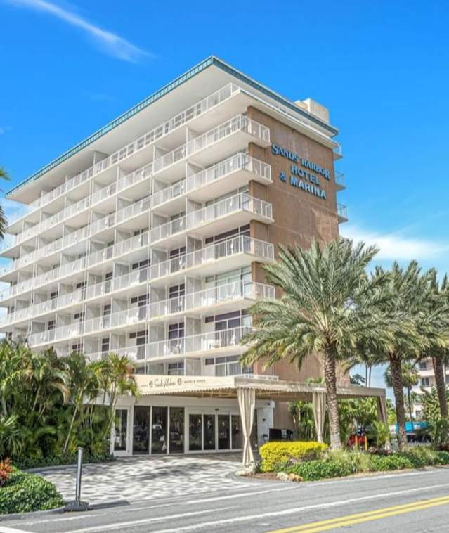 An eight-story hotel with a portico in front. Several palm trees and greenery line the drive